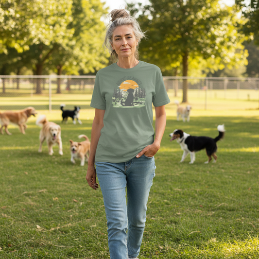 A woman wearing a sage green graphic t-shirt with a dog silhouette and sunset print, paired with blue jeans, set against a plain white background.